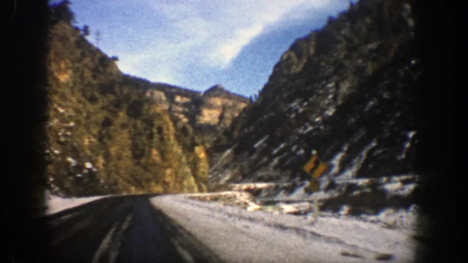 1961 Aspen, Colorado home-movie frame showing a curving mountain road, snowy shoulder, rocky cliffs, and evergreen trees from a moving car.