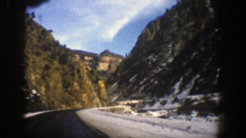 Passenger-view 8mm frame of a Colorado mountain road bending past rocky cliffs, snow patches, evergreen trees, and a pale blue sky.