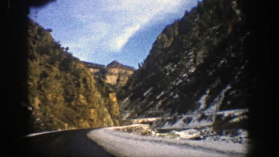 1961 Colorado road-trip footage frame showing a paved road, snow-covered shoulder, steep mountain walls, and evergreen trees from inside a vehicle.