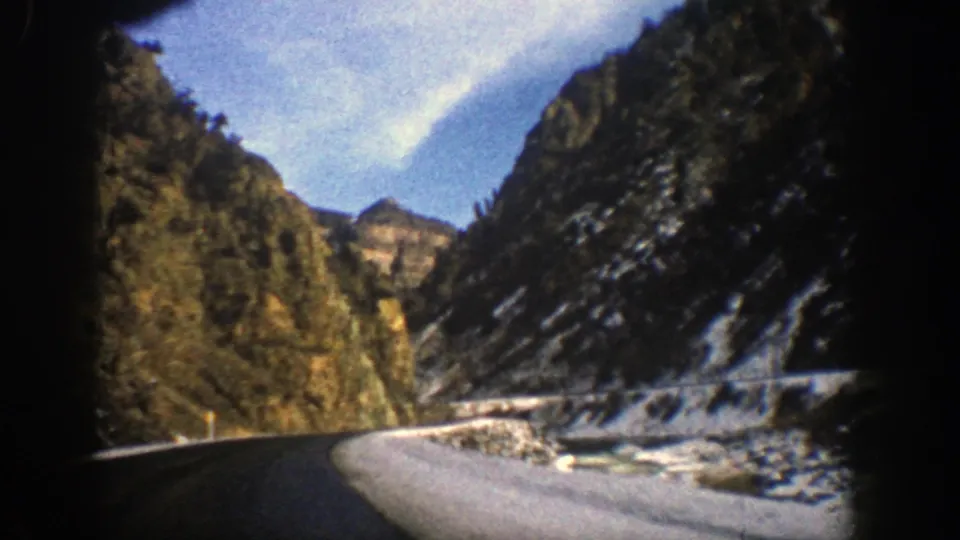Vintage 8mm Aspen, Colorado frame with a winding road, snowy roadside, high rocky slopes, scattered evergreens, and visible film-frame edges.