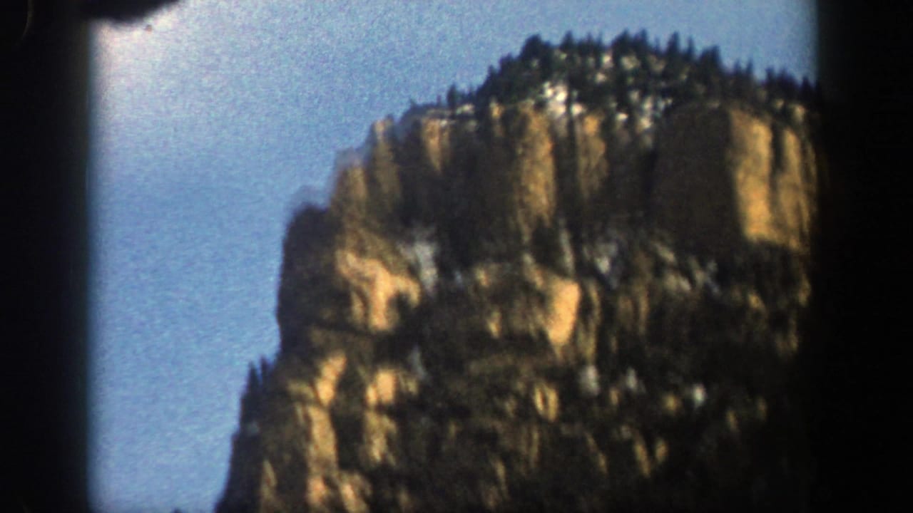 Rocky cliff topped with evergreens against a blue sky.