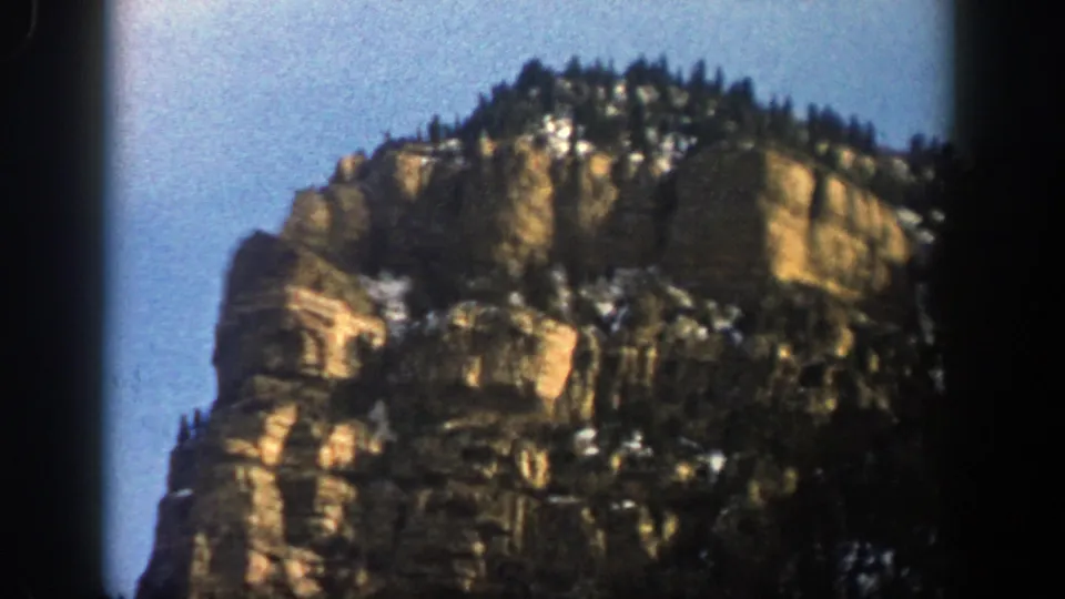 1961 Aspen, Colorado 8mm frame of a rugged cliff face, evergreen trees, and scattered snow under a clear blue sky.