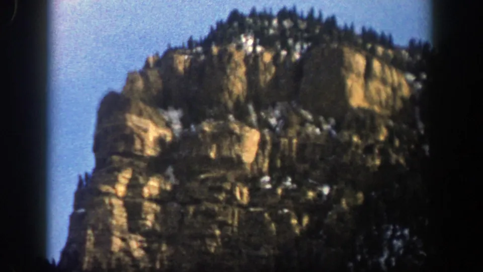 1961 Aspen, Colorado 8mm frame showing layered canyon rock, dark evergreens, and patches of snow on the slope.