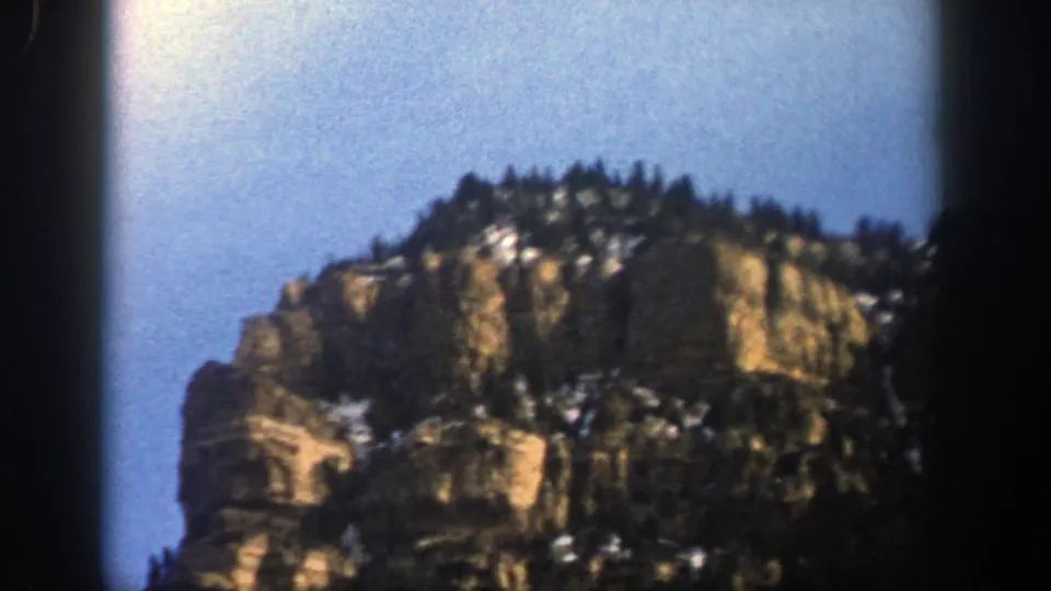 1961 Aspen, Colorado 8mm frame with the upper portion of a rocky cliff, treeline, snow patches, and blue sky.