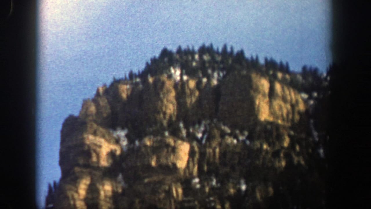 Rocky mountain cliff with evergreens, snow patches, and blue sky.