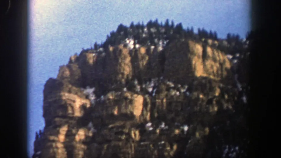 1961 Aspen, Colorado frame of a rugged, snow-dusted rock formation crowned by evergreen trees under clear sky.