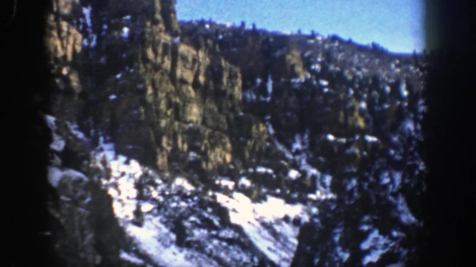 1961 Aspen, Colorado frame showing a steep rocky slope, evergreen trees, and bright snowfields in an 8mm scan.
