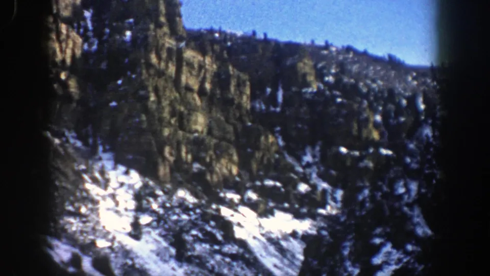 1961 Aspen, Colorado 8mm frame with a snow-streaked rocky ridge, evergreen forest, and clear blue sky.