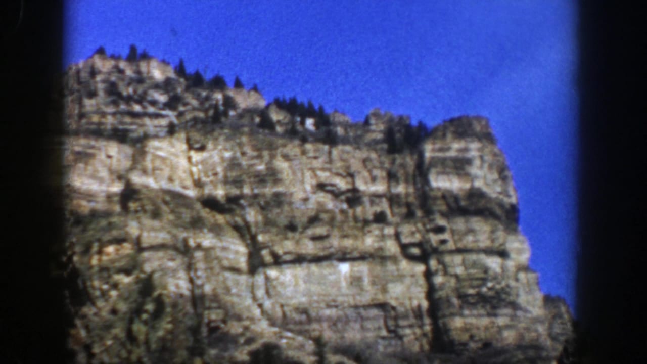 1961 Aspen, Colorado home-movie frame showing a layered rocky mountain face, ridge-top evergreens, and clear blue sky.