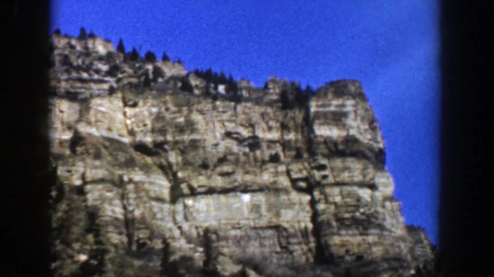 1961 Aspen, Colorado frame of a rugged stratified mountain wall with sparse evergreens silhouetted against blue sky.