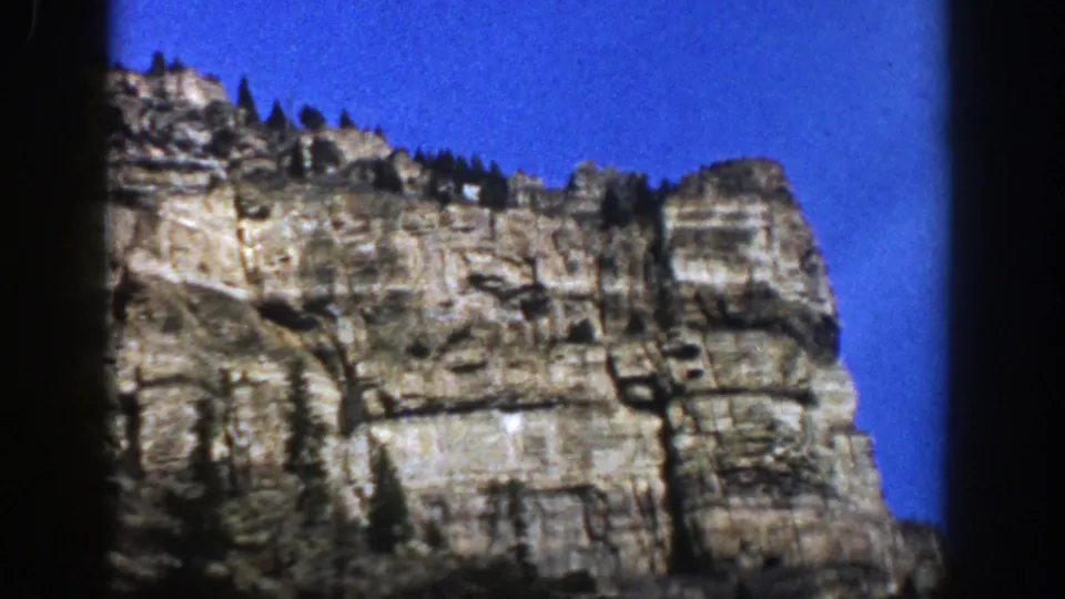 1961 Aspen, Colorado frame showing a rocky cliff, dark ridge trees, saturated blue sky, and visible 8mm frame borders.