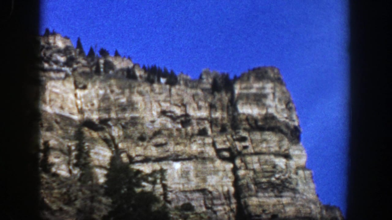 1961 Aspen, Colorado 8mm frame showing a sunlit rock cliff with evergreens along the ridge and dark gate borders.