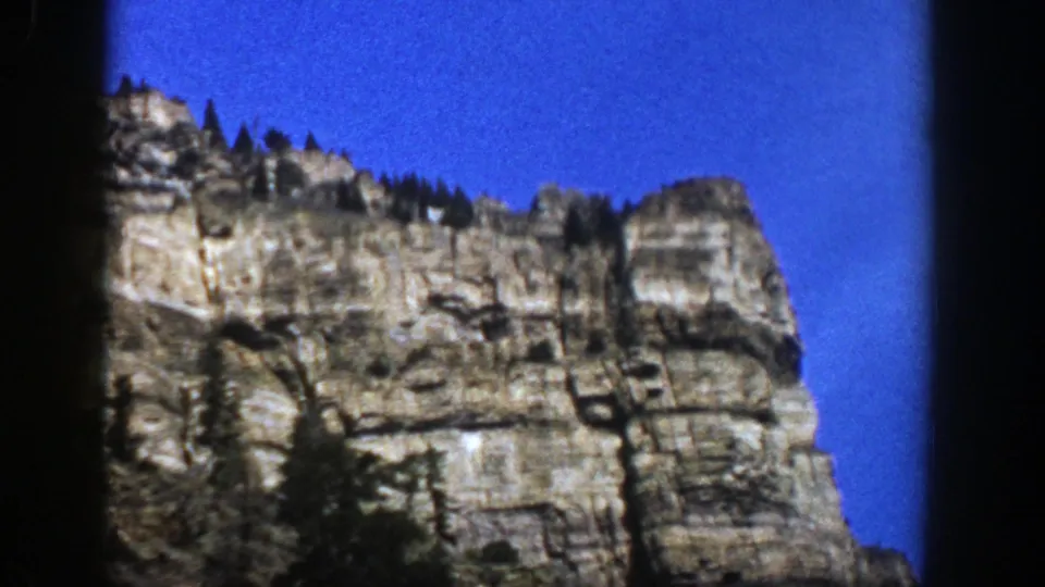 1961 Aspen, Colorado 8mm frame showing a sunlit rock cliff with evergreens along the ridge and dark gate borders.
