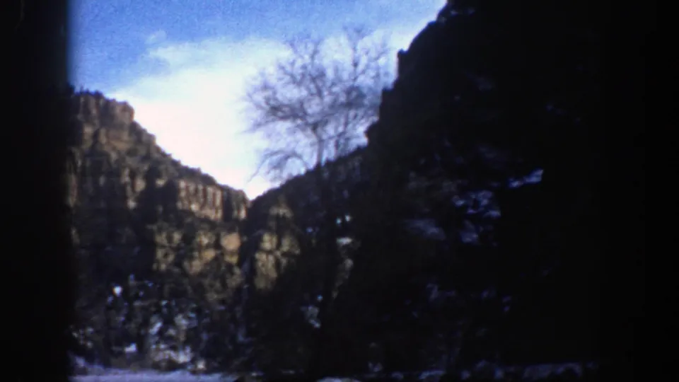 1961 Aspen, Colorado canyon view with rocky slopes, snow patches, dark evergreens, and a leafless tree against pale sky.