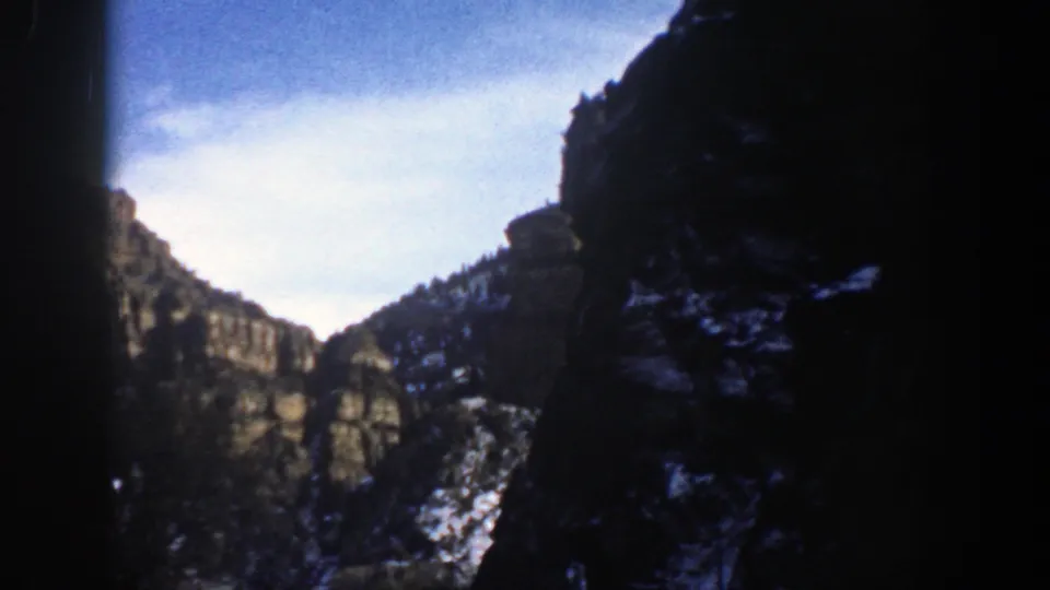 1961 Aspen, Colorado 8mm frame looking through a shadowed canyon toward rocky cliffs, snow patches, trees, and bright sky.