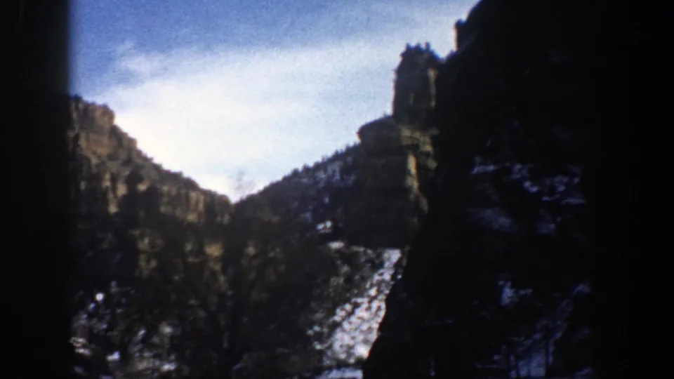 1961 Aspen, Colorado frame showing a rocky slope and cliff tower with snow patches, dark evergreens, pale sky, and heavy film-edge shadow.