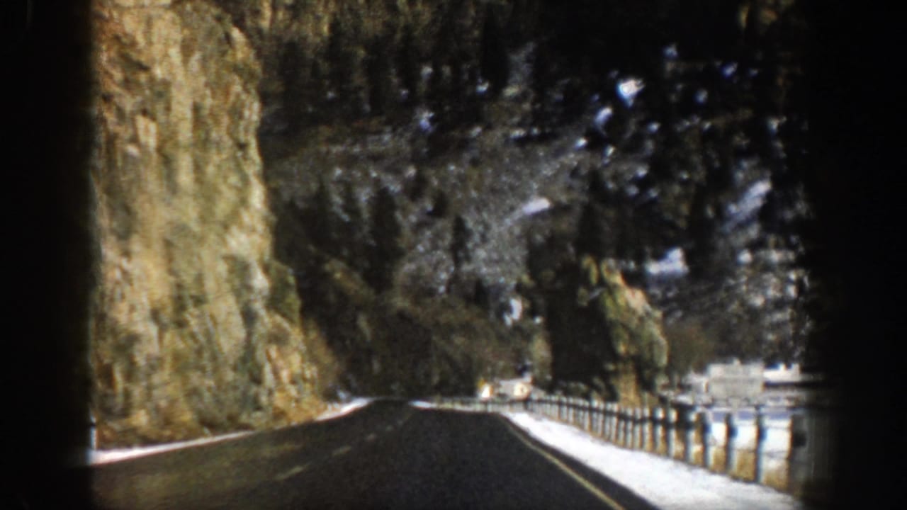 Wet mountain road beside rocky cliffs with snow along the shoulder.