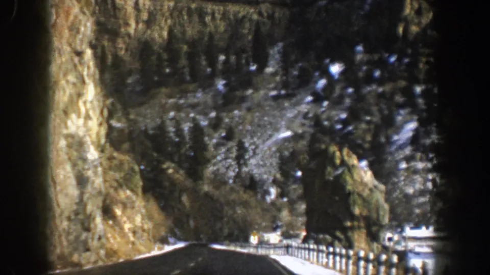 Passenger-view frame from 1961 Aspen footage with a dark road, snow patches, rocky canyon walls, guardrail posts, and forested slopes.
