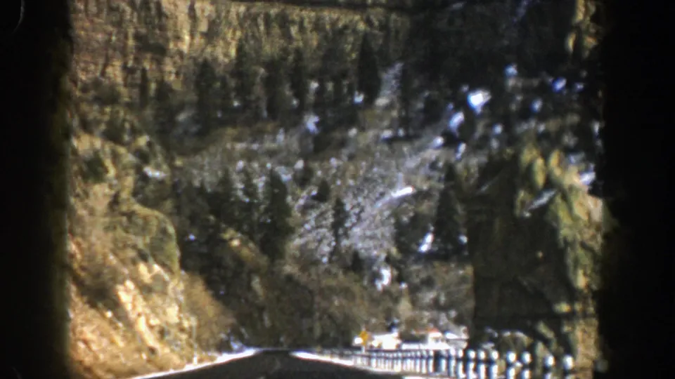 1961 Aspen, Colorado road-view frame showing a wet lane, cliff face, guardrail, snow, and dark evergreen-covered terrain.