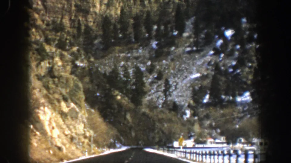 Late frame from the 1961 Aspen drive showing the road continuing through a canyon with snowy edges, guardrails, cliffs, and forested rock slopes.