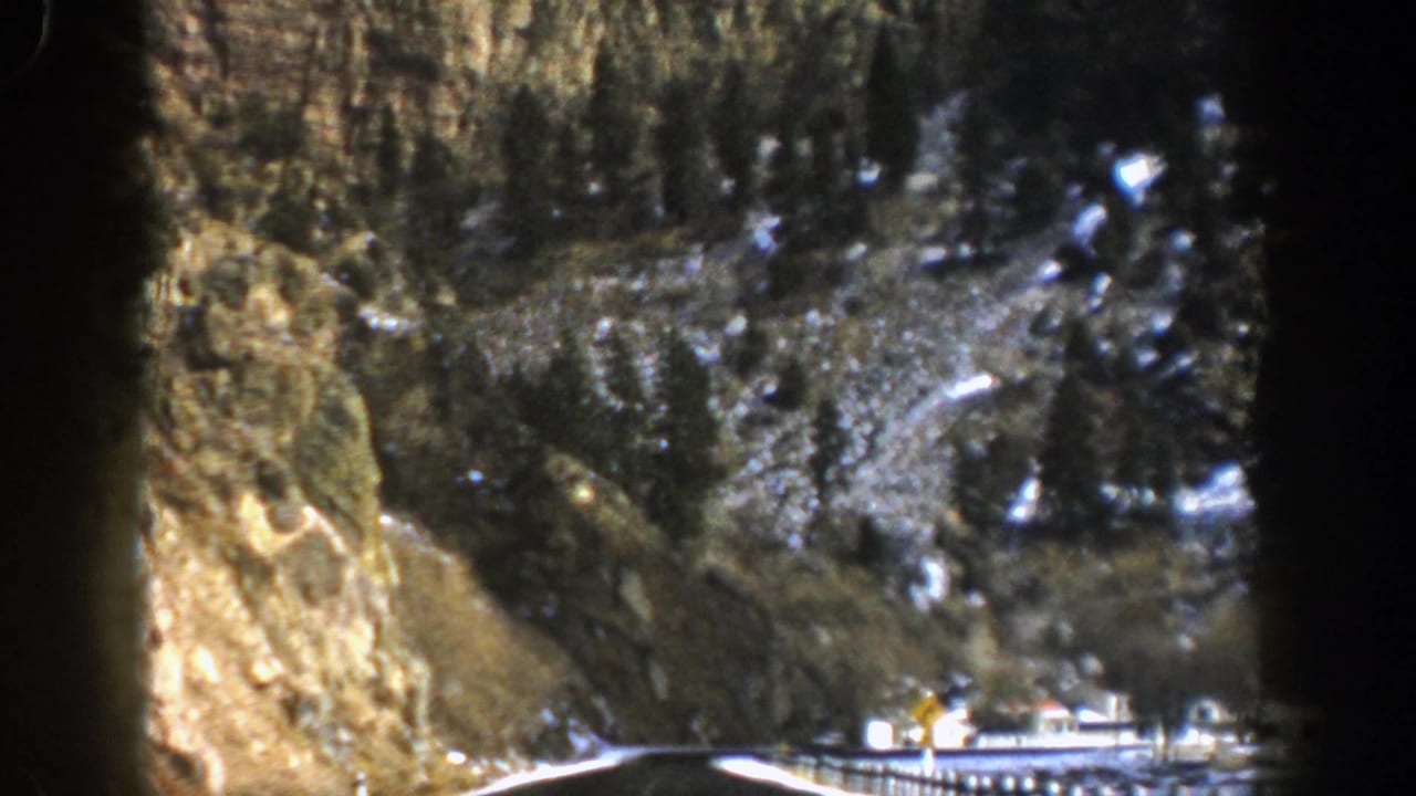 1961 Aspen, Colorado home-movie frame showing a road running between rocky cliffs, evergreen trees, and scattered snow.