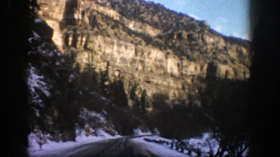 1961 home-movie frame from Aspen, Colorado showing a curving road, roadside snow, guardrail posts, and sunlit cliffs.