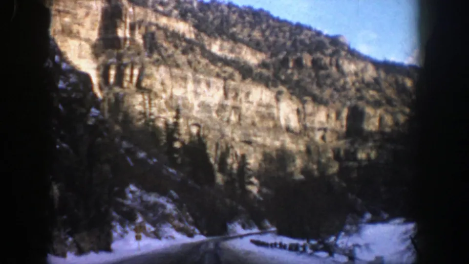 Late-frame 8mm view of a snowy Aspen canyon drive with a road, guardrail, evergreen trees, and layered rock cliffs.