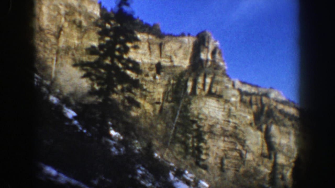 1961 Aspen, Colorado home-movie frame looking upward at a layered cliff with evergreens, snow patches, and clear blue sky.