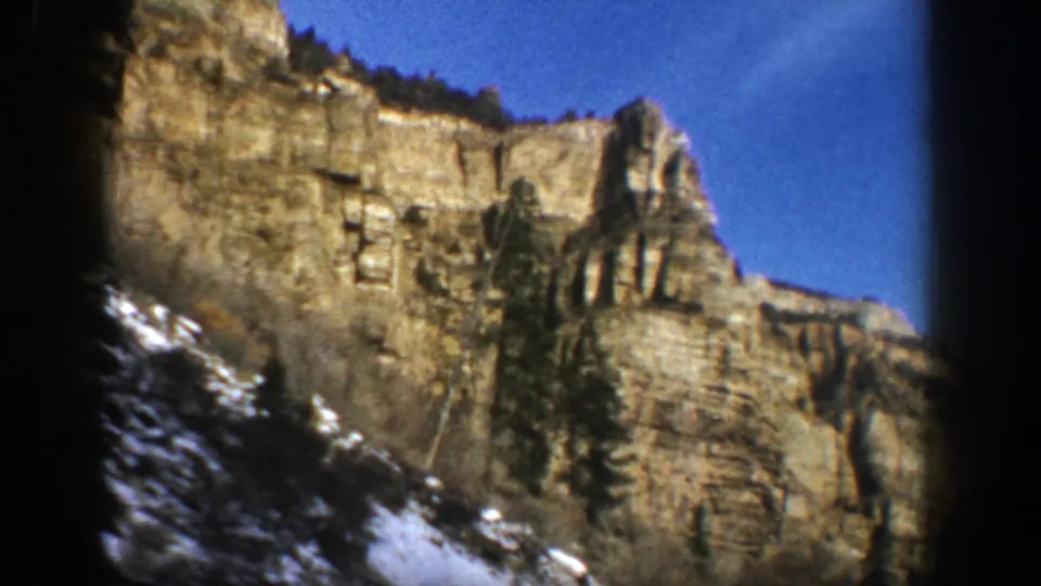 1961 Aspen, Colorado frame showing a tan cliff face rising above evergreens and snow along the lower slope.
