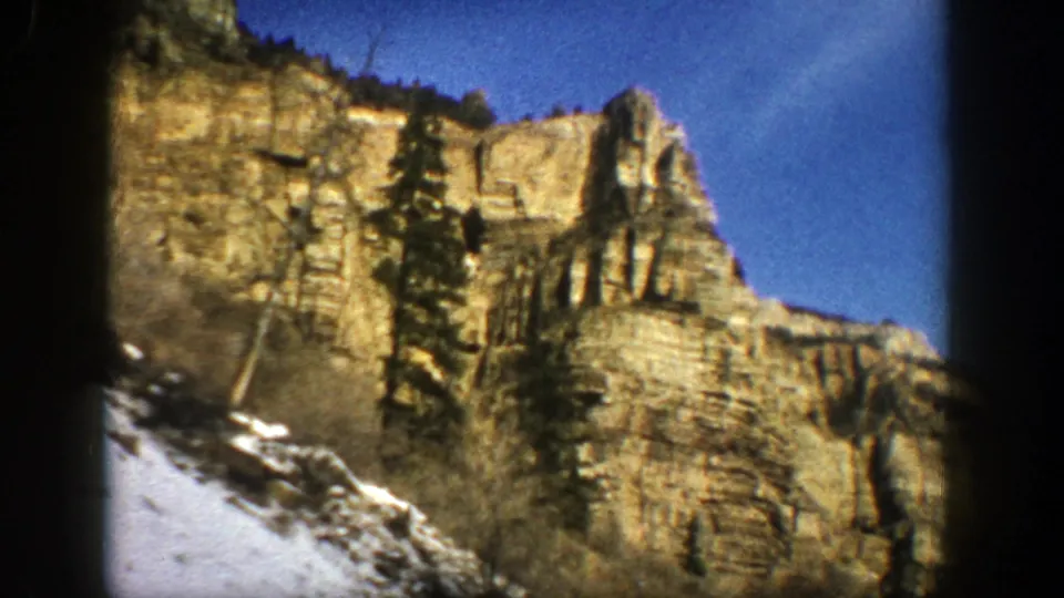 1961 Aspen, Colorado 8mm frame with a tall evergreen in front of layered cliff rock and snow along the bottom edge.