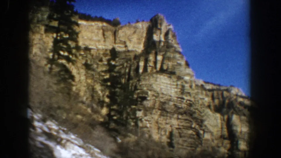 1961 Aspen, Colorado home-movie frame of a cliff and evergreen slope under a deep blue sky, with snow visible near the base.