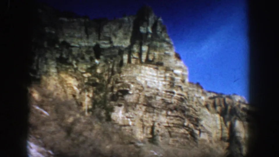 1961 Aspen, Colorado view of a rugged cliff face with shadowed trees, snow on the lower slope, and strong blue sky.
