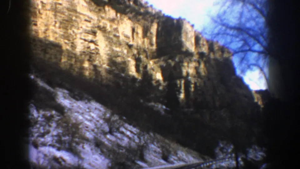 1961 Aspen, Colorado mountain scene with sunlit layered rock, snow patches, sparse trees, and bare branches at frame edge.