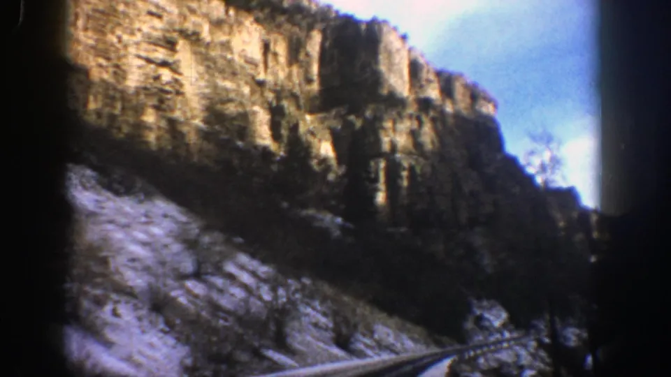 1961 Aspen-area 8mm frame showing a rocky cliff face, snow-dusted roadside slope, and a road curving through a mountain canyon.