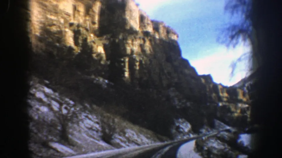 1961 Aspen-area 8mm frame with a winding road, high rock formations, snow patches, and dark film borders from the moving-car viewpoint.