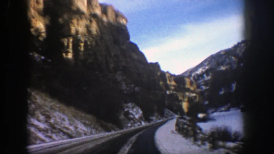 1961 Aspen-area 8mm frame looking down a canyon road toward cliffs and a bright snow or water surface beside the route.