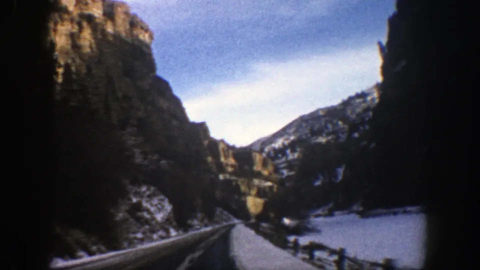 1961 Aspen-area 8mm frame showing a road curving past dark cliffs, snow-covered slopes, and a bright surface at the canyon floor.