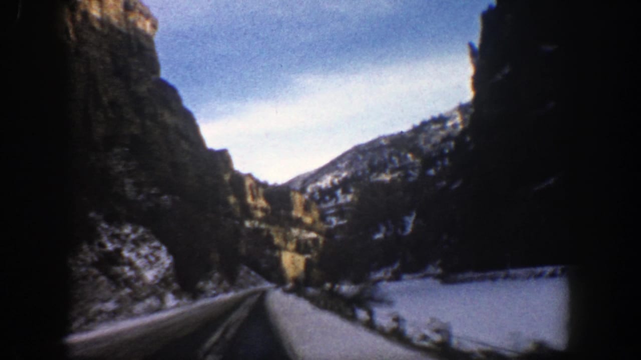 1961 Aspen-area 8mm view from a car on a snow-covered mountain road between steep cliffs and winter slopes.