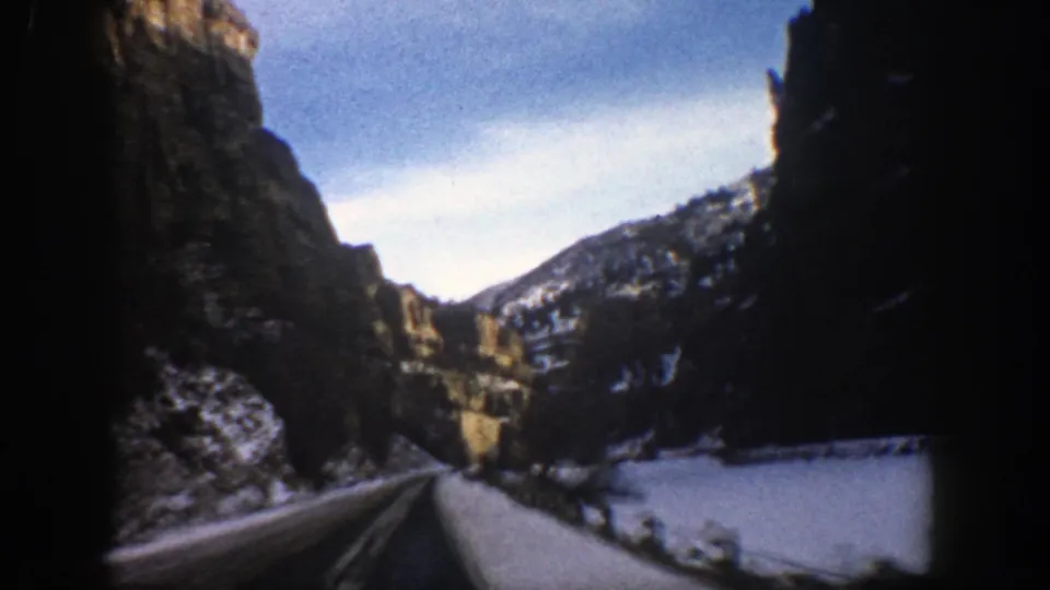 1961 Aspen-area 8mm view from a car on a snow-covered mountain road between steep cliffs and winter slopes.