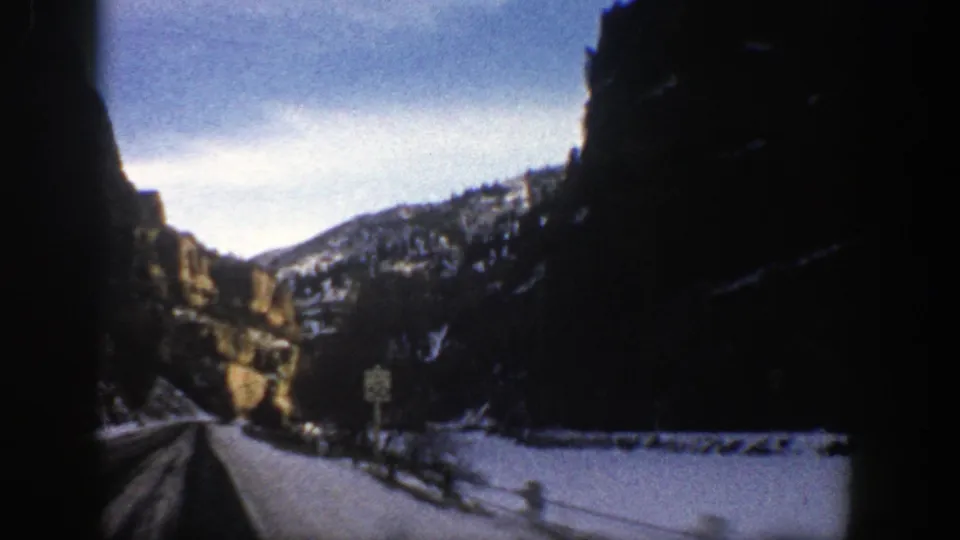 1961 Aspen, Colorado home-movie frame with a snowy roadside, cliff shadows, a small sign, and distant winter mountains.