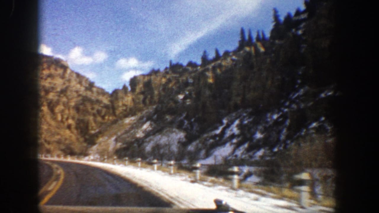 Curving mountain road beside snowy shoulder and rocky slopes under blue sky.
