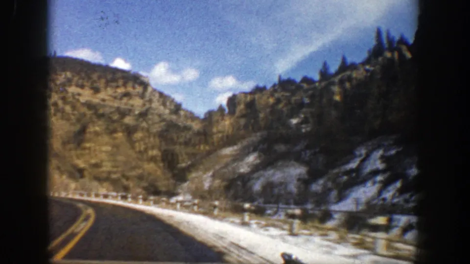 1961 Aspen, Colorado frame from a moving vehicle, with a paved road, snow patches, rocky brown slopes, evergreens, and bright sky.
