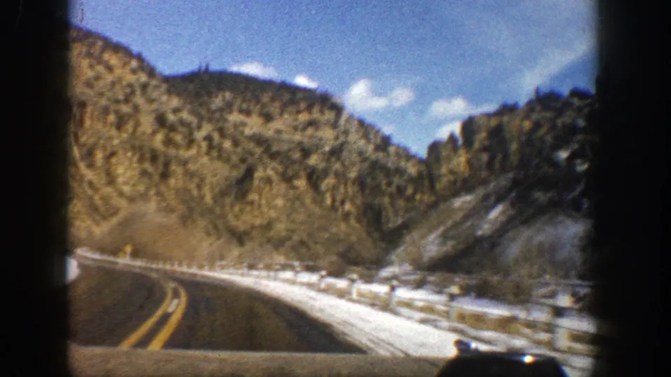 1961 Aspen, Colorado home-movie frame showing the road edge, snow-covered shoulder, rocky hillside, and trees from a moving car.