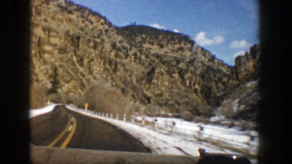 1961 Aspen, Colorado 8mm frame of a snow-lined two-lane road approaching brown rocky mountains and scattered evergreen trees.
