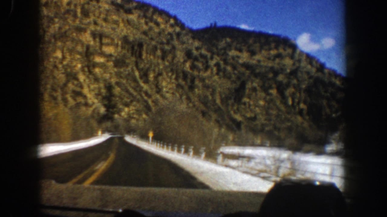 1961 Aspen-area home-movie frame showing a forward view along a paved mountain road bordered by snow and steep rocky terrain.