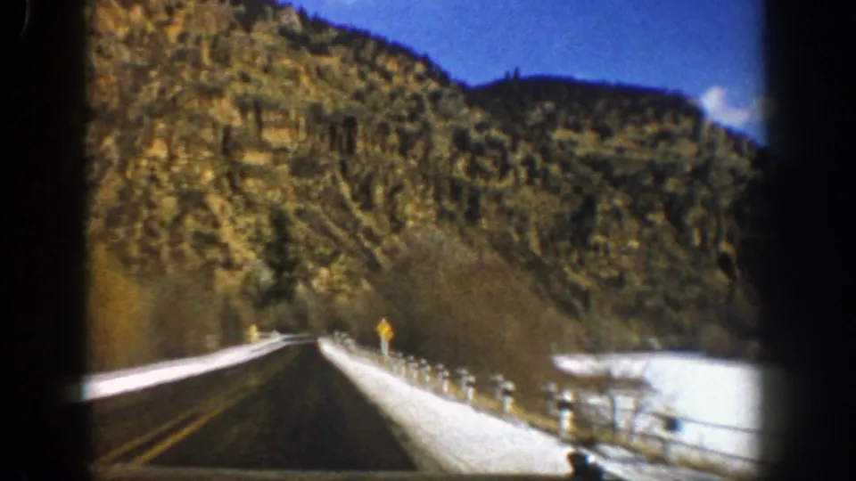 1961 Aspen-area road footage frame with a paved roadway, snow-covered roadside, guard posts, and rugged hills in winter light.