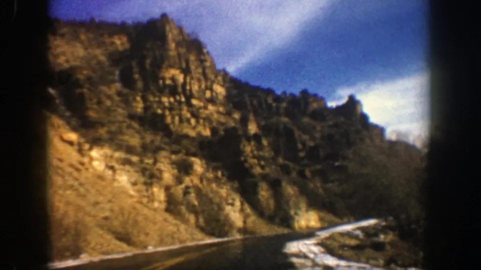 1961 Aspen-area home-movie frame showing the road bending past steep tan rock formations and snow patches in a mountain valley.