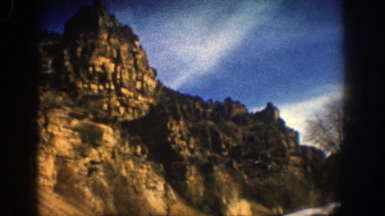 1961 Aspen, Colorado home-movie frame showing rugged cliffs above a road with snow patches and blue sky.