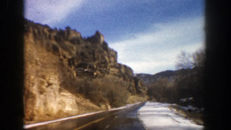 Vintage 8mm view of an empty Aspen-area mountain road bordered by cliffs, winter brush, and snow.