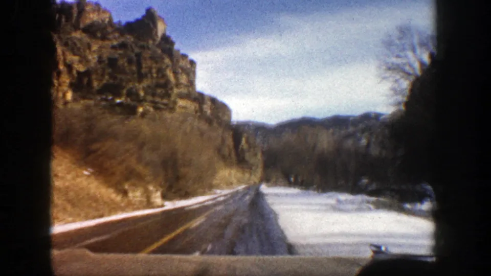 1961 Aspen, Colorado driving view with a cliff wall, snowy roadside, bare trees, and distant hills ahead.